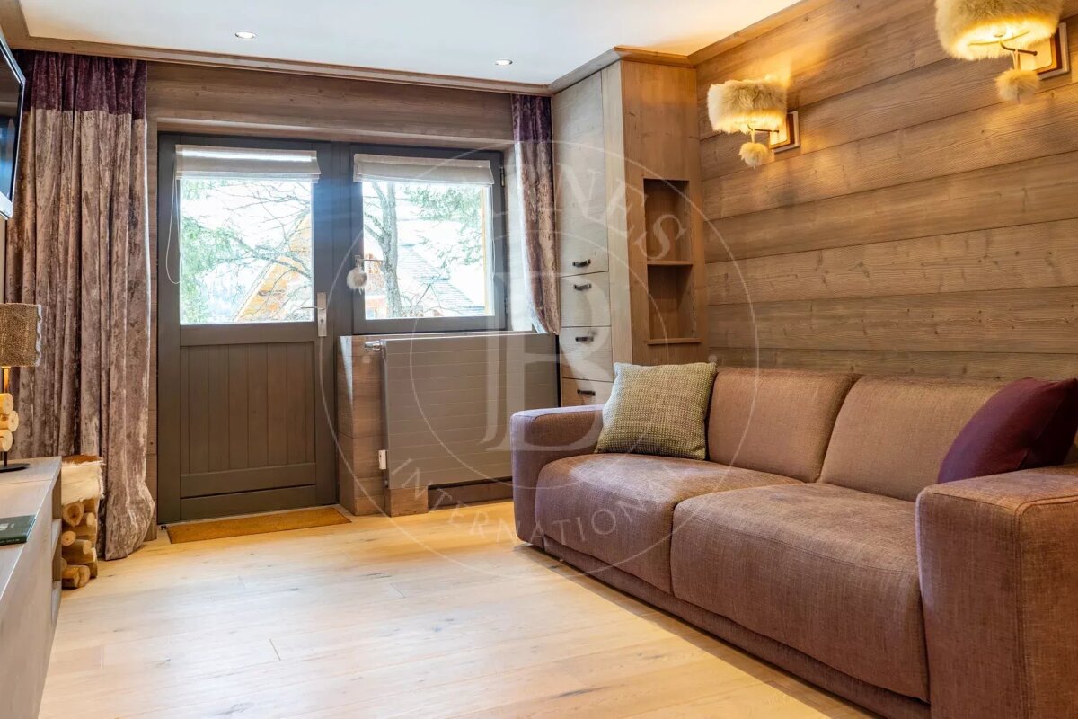 A cozy living room features light wooden walls and floors, a large brown sofa, and a window/door offering a view of trees. Furry wall lights add warmth.