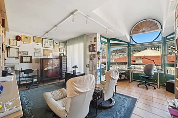 A well-lit, eclectic living room/study with walls full of framed items, comfortable recliners, and a wide window offering a scenic city view.