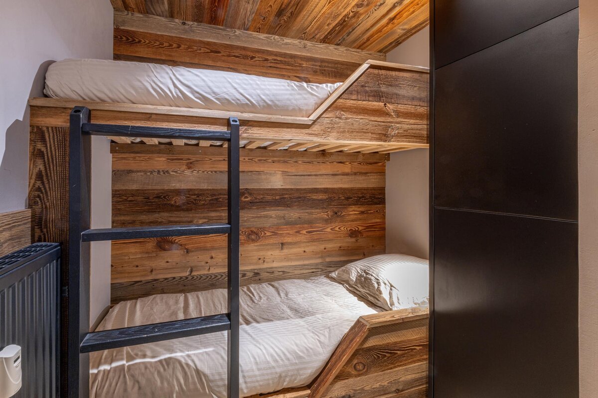 Cozy room with rustic wooden bunk beds and white bedding. Features a black ladder, wood-paneled walls/ceiling, and a dark wardrobe.