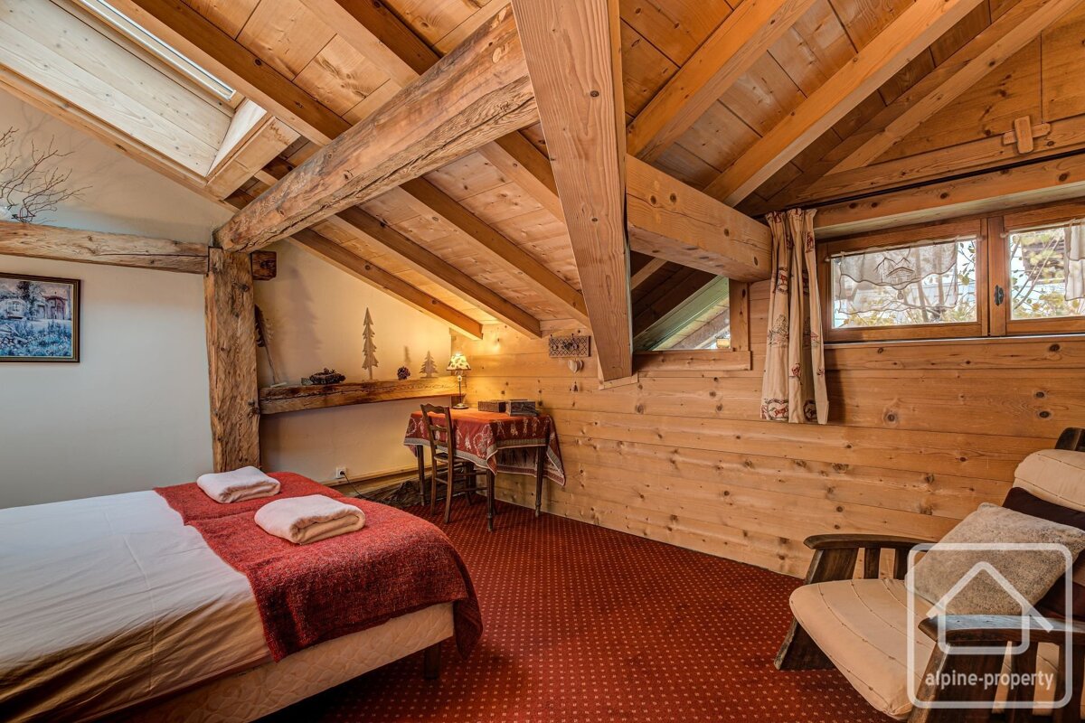 Rustic attic bedroom with wood-paneled walls and ceiling, exposed beams, a bed with red blanket, and a red patterned carpet.