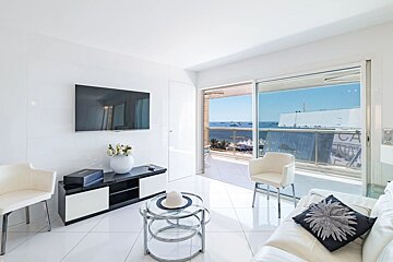 Minimalist white living room with sleek furniture, a TV, and a balcony offering a wide view of the blue sea with distant boats.