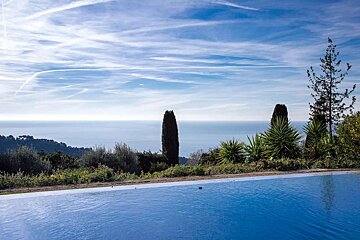 An infinity pool overlooks a vast blue ocean and a green hillside, under a blue sky streaked with white contrails.