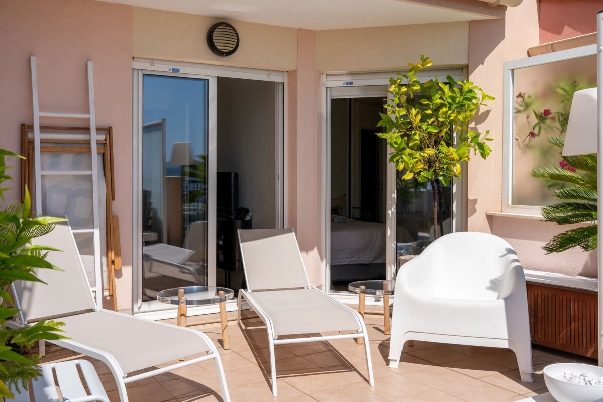 Sunny tiled terrace with beige loungers, a white armchair, potted plants, and glass sliding doors.