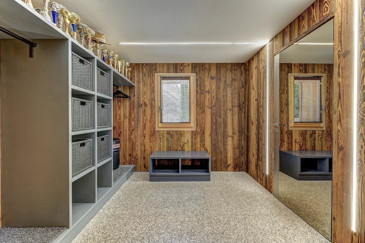 Cozy rustic room with wood-paneled walls, carpet, trophy shelf, grey storage, bench, window, and mirror with LED strips.