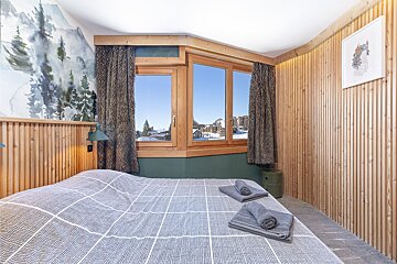 Cozy chalet bedroom featuring wood paneling, a large bed with grey towels, and a window overlooking snowy mountain views.