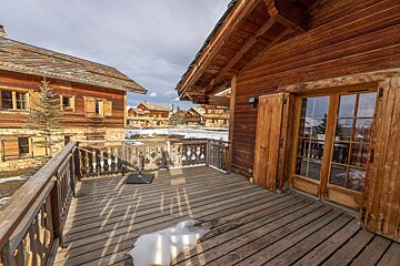 A rustic wooden chalet balcony with snow patches, overlooking a snowy mountain village with other chalets under a cloudy sky.