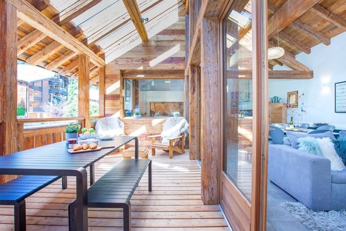 Sunlit wooden chalet terrace with an outdoor dining set under a glass ceiling, beside a cozy, modern living room.