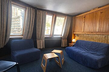 A cozy room with blue carpet, a large blue sofa, and chairs. Wooden wall paneling, built-in storage, and slanted windows with beige curtains complete the space.