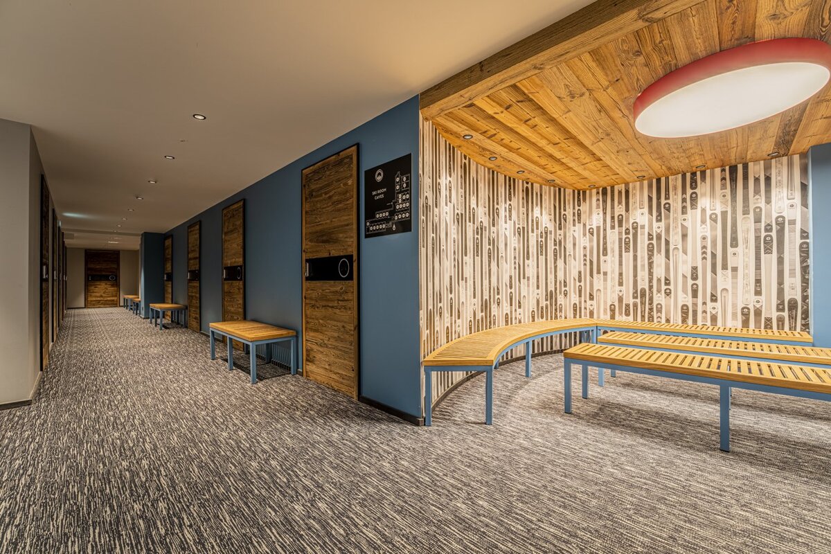 Modern ski lodge corridor with blue walls, wooden doors, patterned carpet, and a waiting area featuring wooden benches, ski-patterned wallpaper, and a wooden ceiling.