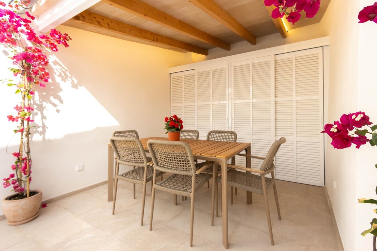 A bright outdoor patio features a wooden dining table with six woven chairs, vibrant pink bougainvillea, white louvered doors, and a beamed ceiling.