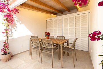 A bright outdoor patio features a wooden dining table with six woven chairs, vibrant pink bougainvillea, white louvered doors, and a beamed ceiling.