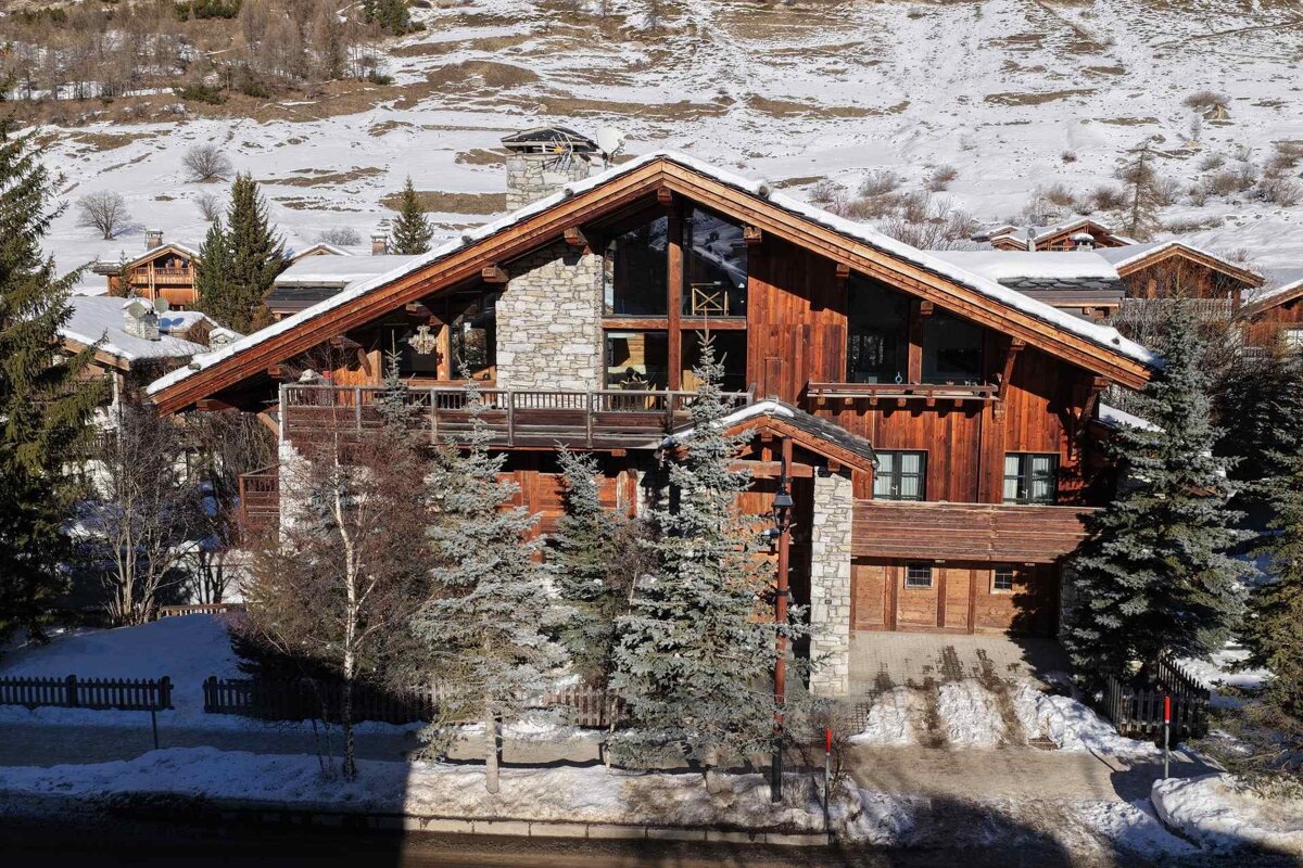 A luxurious wooden and stone chalet with a snow-covered roof and towering windows, nestled among pine trees in a snowy mountain village.