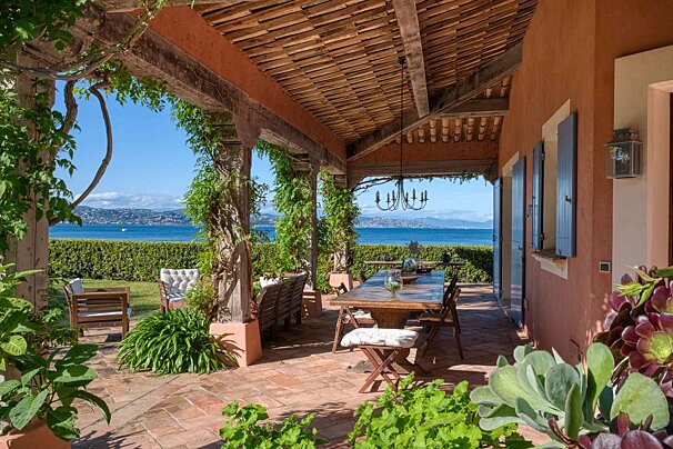 Sunny patio with a vine-covered pergola, outdoor dining, and an expansive view of the calm blue sea and distant coastline.