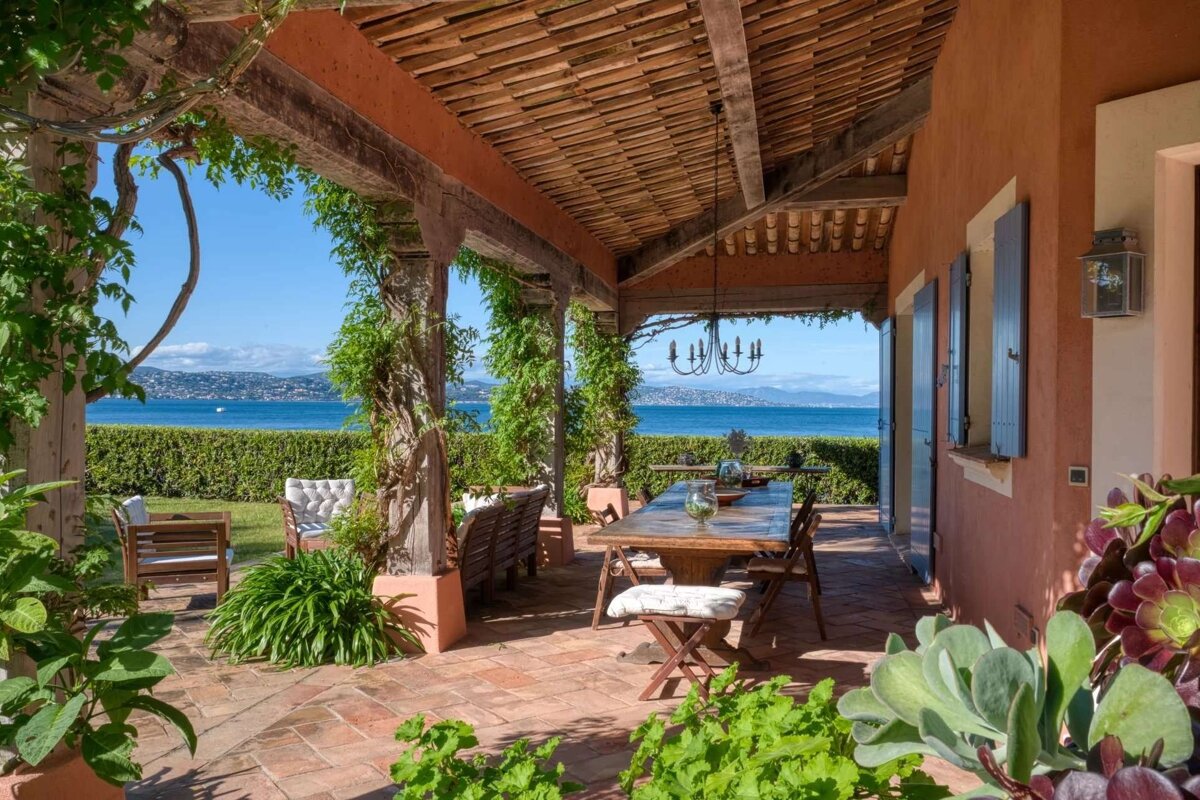 Sunny patio with a vine-covered pergola, outdoor dining, and an expansive view of the calm blue sea and distant coastline.