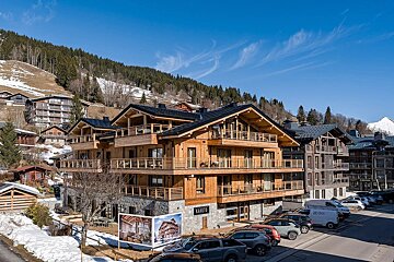 A large wooden alpine chalet named "Kangto" in a mountain ski resort with parked cars under a clear blue sky.