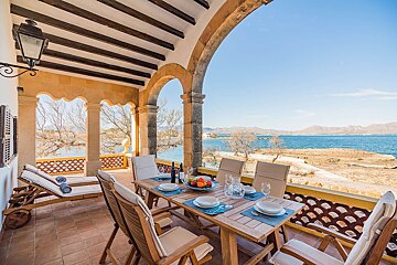 An arched terrace with a set dining table and lounge chairs overlooks a serene blue sea, rocky coast, and distant mountains under a clear sky.