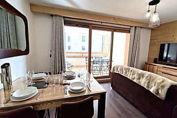 A modern open-plan living and dining area with a set table, cozy sofa, TV, and large glass doors opening to a balcony with outdoor furniture.