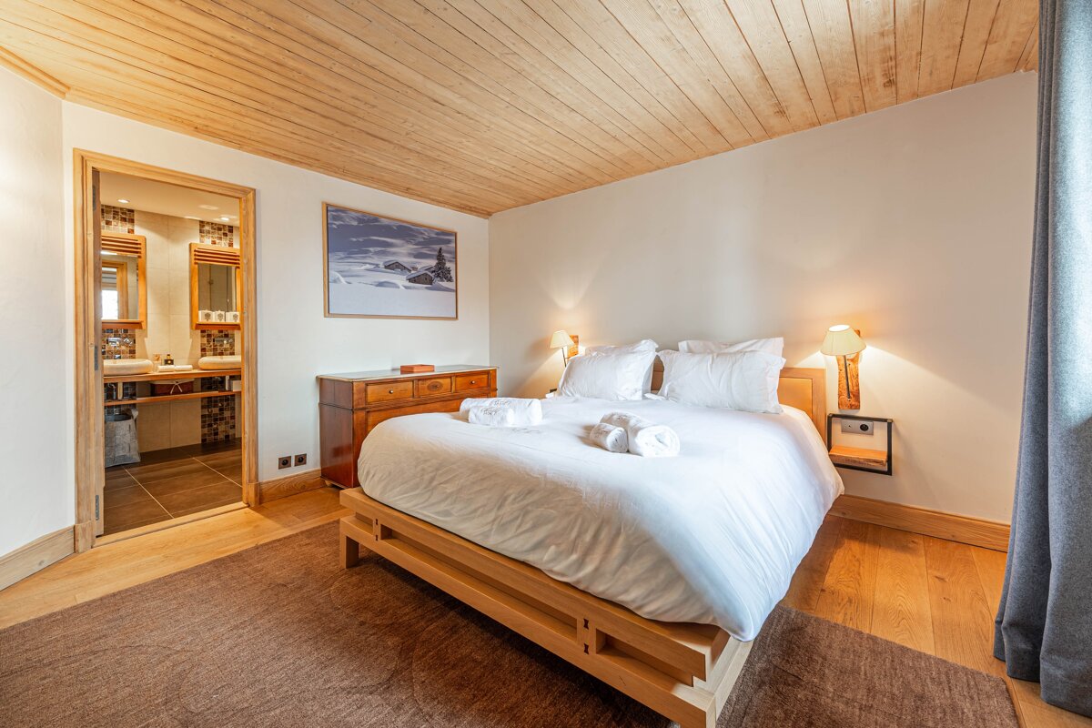 A cozy bedroom with a wooden plank ceiling, white bed, wooden dresser, and an open doorway leading to a tiled bathroom.