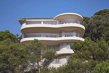 A multi-story, curved white building with glass balconies, nestled among lush green trees against a clear blue sky.