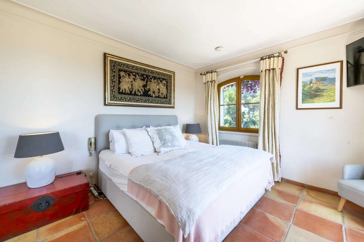 A cozy bedroom with a bed, white linens, terracotta floor, a window, and varied wall art.
