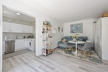 Bright, modern open-plan apartment featuring a white kitchen, blue sofa living area, small dining table, and light wood flooring.
