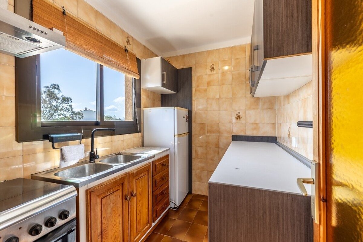 Narrow kitchen with wooden cabinets, a sink, tiled walls, and a white refrigerator under a bright window.