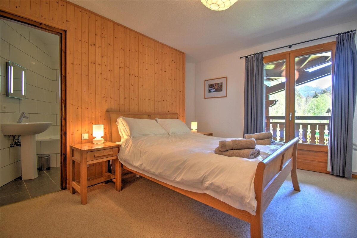A cozy bedroom with wooden paneling, a double bed, and a balcony offering mountain views. An open door reveals a bathroom with a sink.