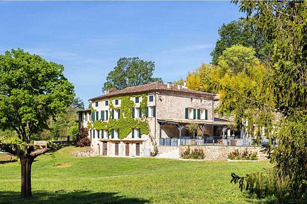 A large stone house with green shutters is surrounded by trees
