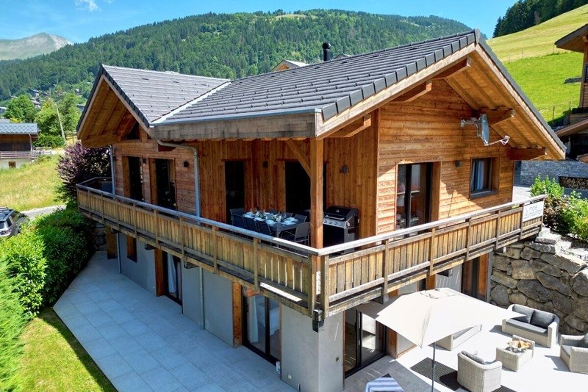 A two-story wooden mountain chalet with a wrap-around balcony, lower patio, and scenic green hills in the background.