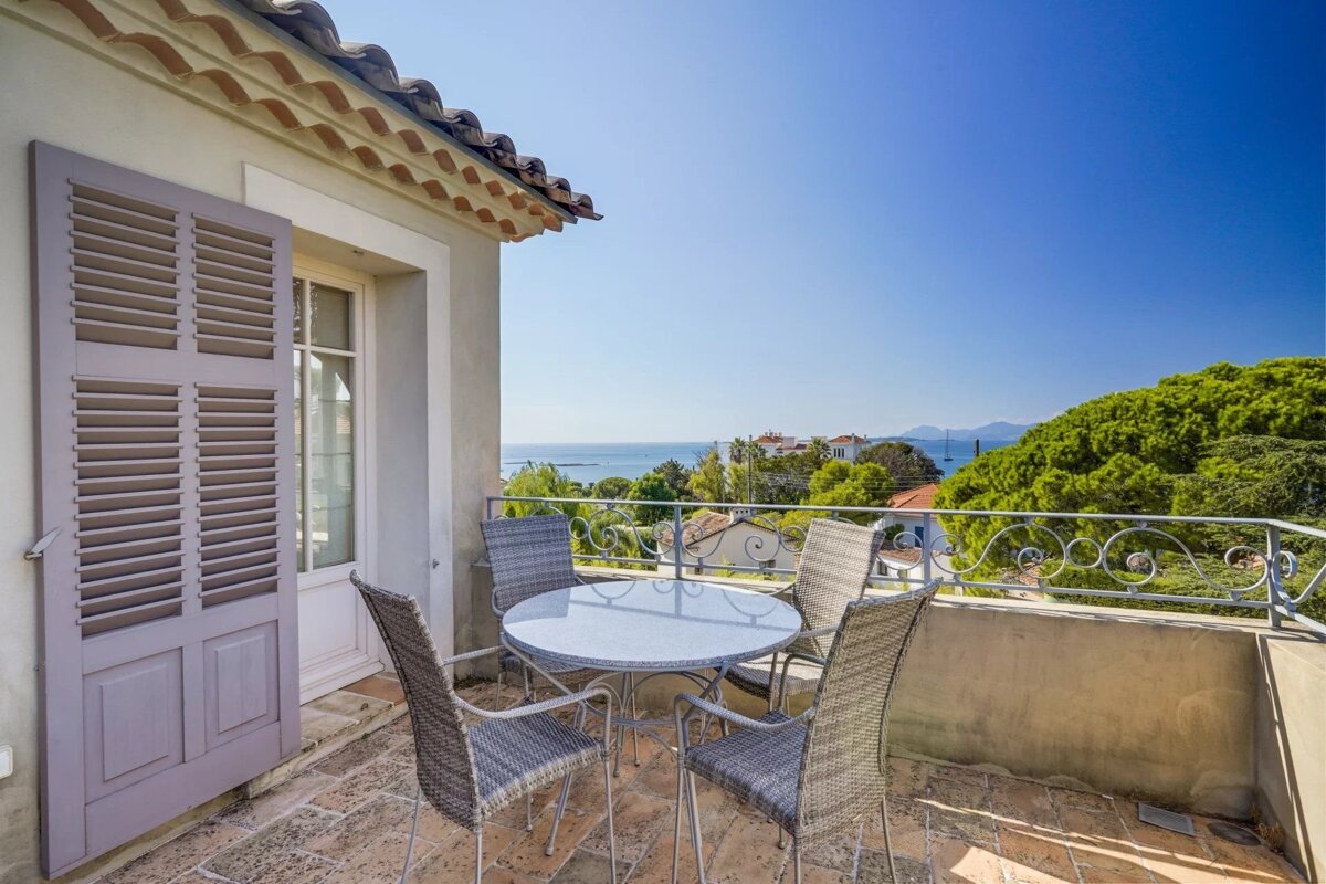 A sunny terrace with a table and four chairs overlooks lush trees and the distant sea under a clear blue sky.