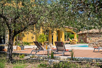 A swimming pool surrounded by chairs and trees with a yellow house in the background