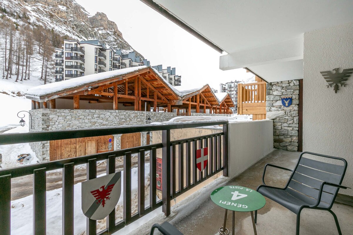 A balcony with a chair and green table overlooks a snowy mountain resort, featuring traditional wooden chalets, modern buildings, and a rugged peak.