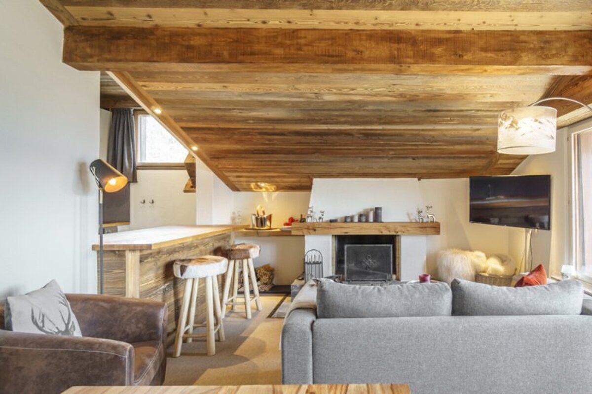 Cozy alpine chalet living room with a low wooden ceiling, fireplace, grey sofa, and rustic bar counter with stools.