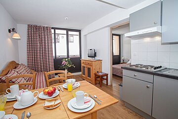A cozy apartment interior featuring a dining table set for breakfast, a kitchenette, and a bedroom visible. A window overlooks a snowy scene.