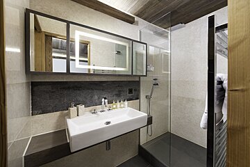 A modern bathroom featuring a white rectangular sink, an illuminated mirror, and a glass shower. It has stone tiles and wooden accents.