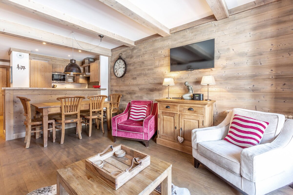 Charming chalet-style living space with wood-paneled walls, wooden kitchen/dining area, two armchairs (pink, white), and a TV.