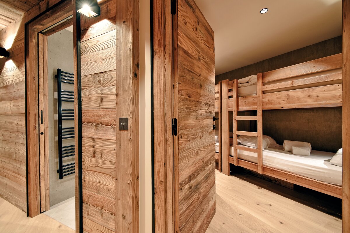 Modern wooden chalet interior with an open bathroom door on the left and a bedroom with built-in wooden bunk beds on the right.