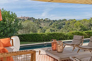 A sunny outdoor patio with a swimming pool, lounge chairs, and orange cushions, overlooking a lush green valley with a distant village under a blue sky.