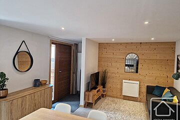 Modern apartment interior with light wood accents, a small living area, a wooden entrance door, and a round hanging mirror.