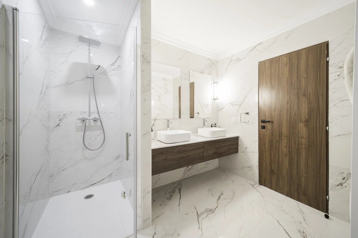 A modern bathroom with white marble walls/floor, glass shower, double vanity with white sinks and wood cabinets, and a wooden door.