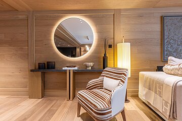 Warm, wood-paneled room featuring a large backlit round mirror, a striped armchair, and cozy glowing lamps.
