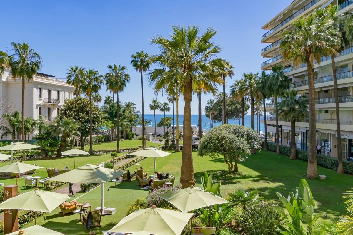 Lush resort garden with palm trees, green umbrellas, and lawn seating with a view of the blue sea in the background.