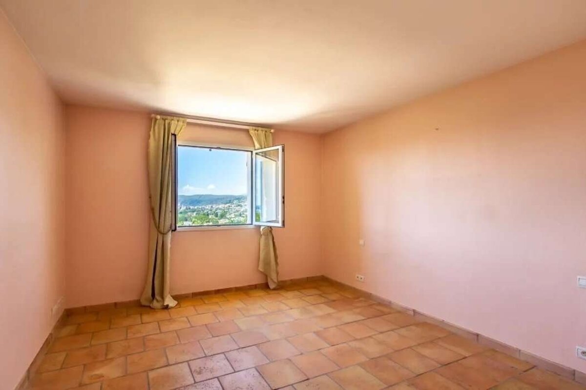 Empty room with pink walls, terracotta floor, and an open window revealing a town view under blue sky. Beige drapes frame the window.