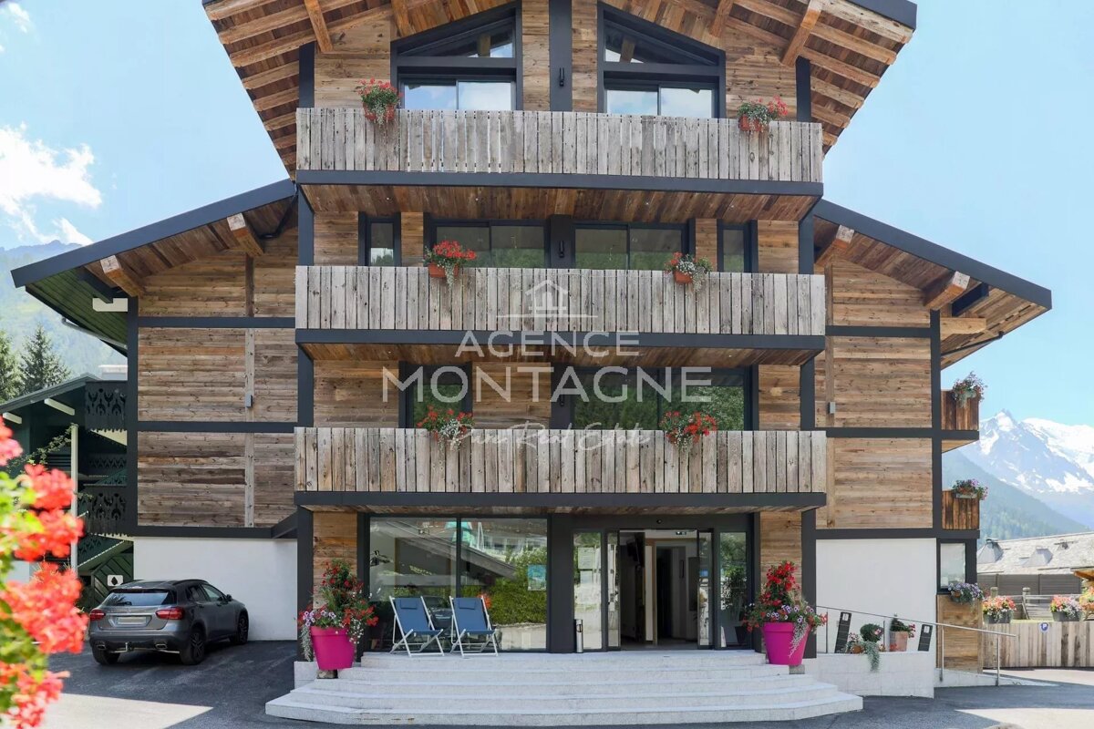 A modern multi-story wooden building with balconies, adorned with flowers. A welcoming entrance features outdoor seating, with mountains in the background.