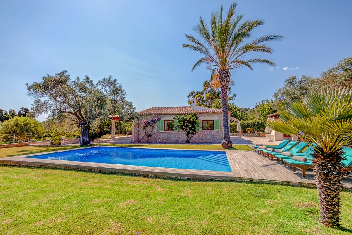 A sunny scene featuring a stone house with green shutters, a sparkling blue swimming pool, lush green lawn, and several palm trees.