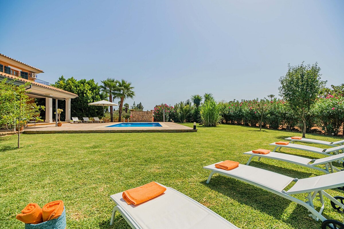 A sunny villa backyard with a green lawn, blue swimming pool, and multiple lounge chairs topped with vibrant orange towels, surrounded by lush greenery.