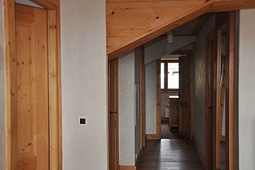 An inviting interior hallway featuring light-colored walls, warm wooden doors, frames, flooring, and angled ceiling beams, leading towards a window.
