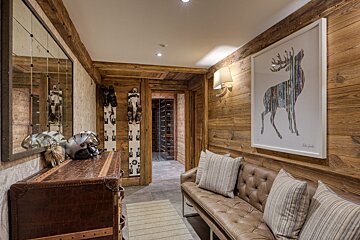 Chic rustic ski lodge entry with wooden walls, displayed skis, helmets, a leather bench, deer art, and a wine cellar beyond.