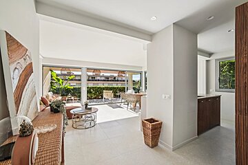 Sunny, modern open-plan apartment with a living room, dining area, and balcony overlooking a hedge and buildings. Wicker decor and light tiles.