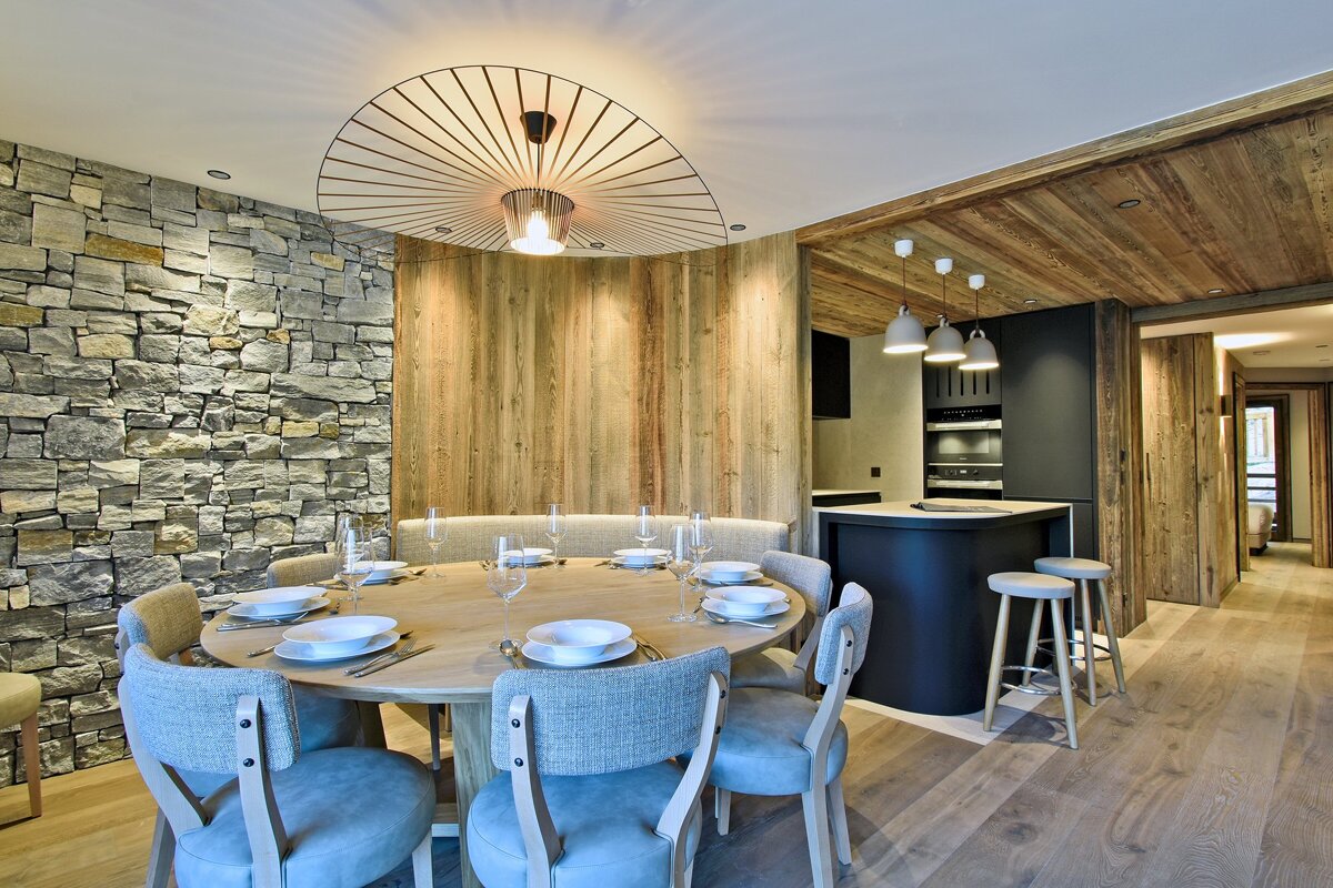 Modern rustic dining area featuring a round table, stone wall, wood paneling, unique ceiling light, and an adjacent sleek kitchen/bar.